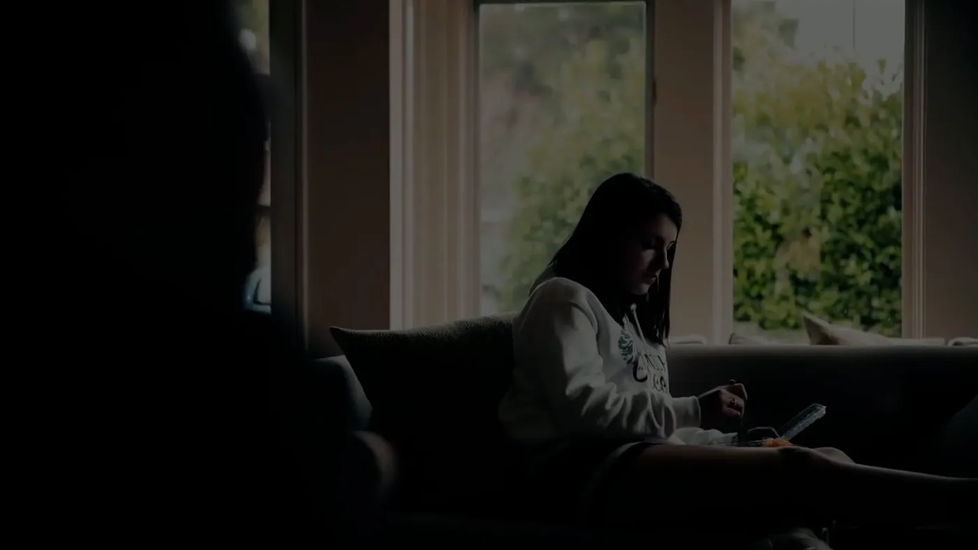 A teenage girl sits alone on a sofa by a bright window, in a dim room.