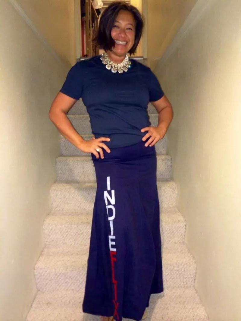 Scilla laughing on a carpeted staircase, hands on hips, in a navy top and a long navy skirt printed with INDIEFLIX down the side.