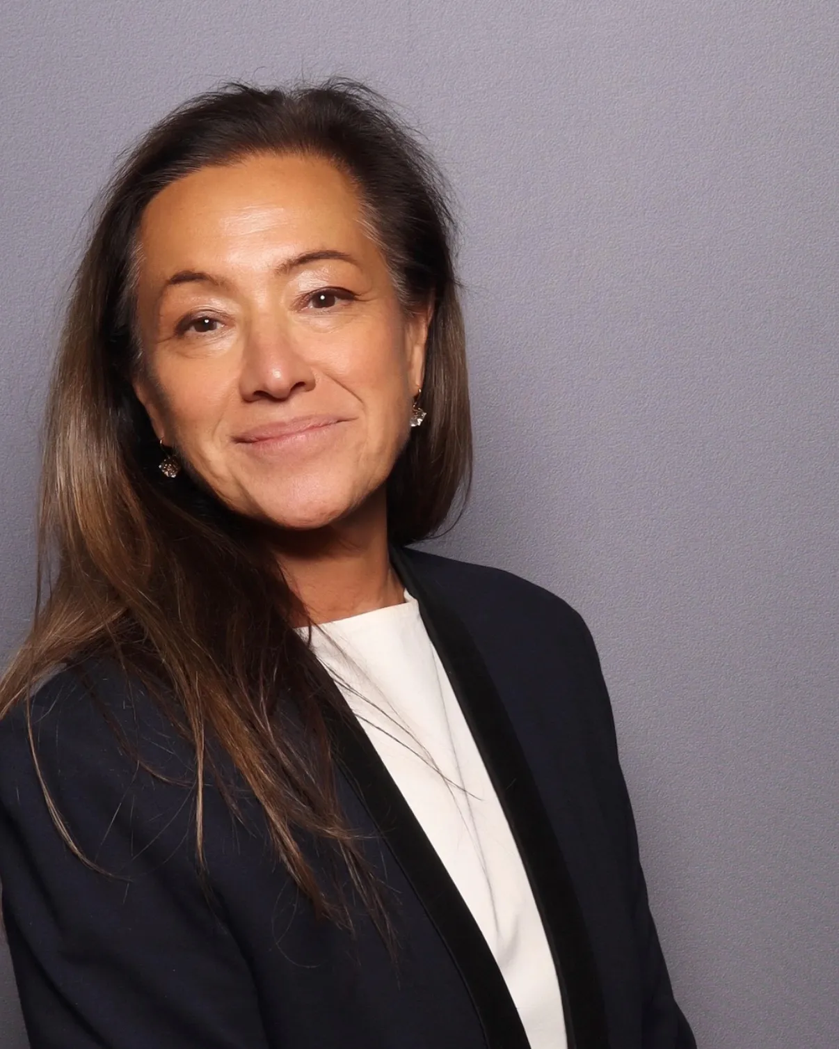 Scilla Andreen in a black blazer over a white top, with a quiet smile, against a soft grey backdrop.