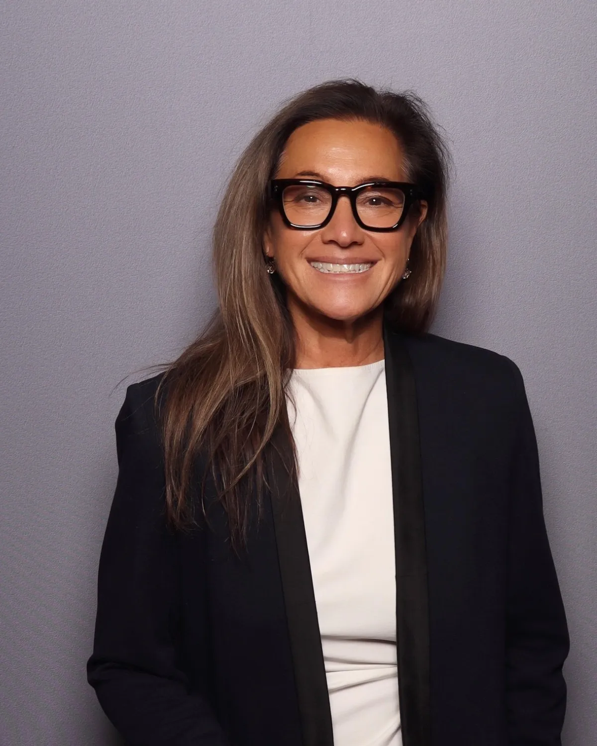 Scilla Andreen smiling in black-framed glasses and a black blazer, against a soft grey backdrop.